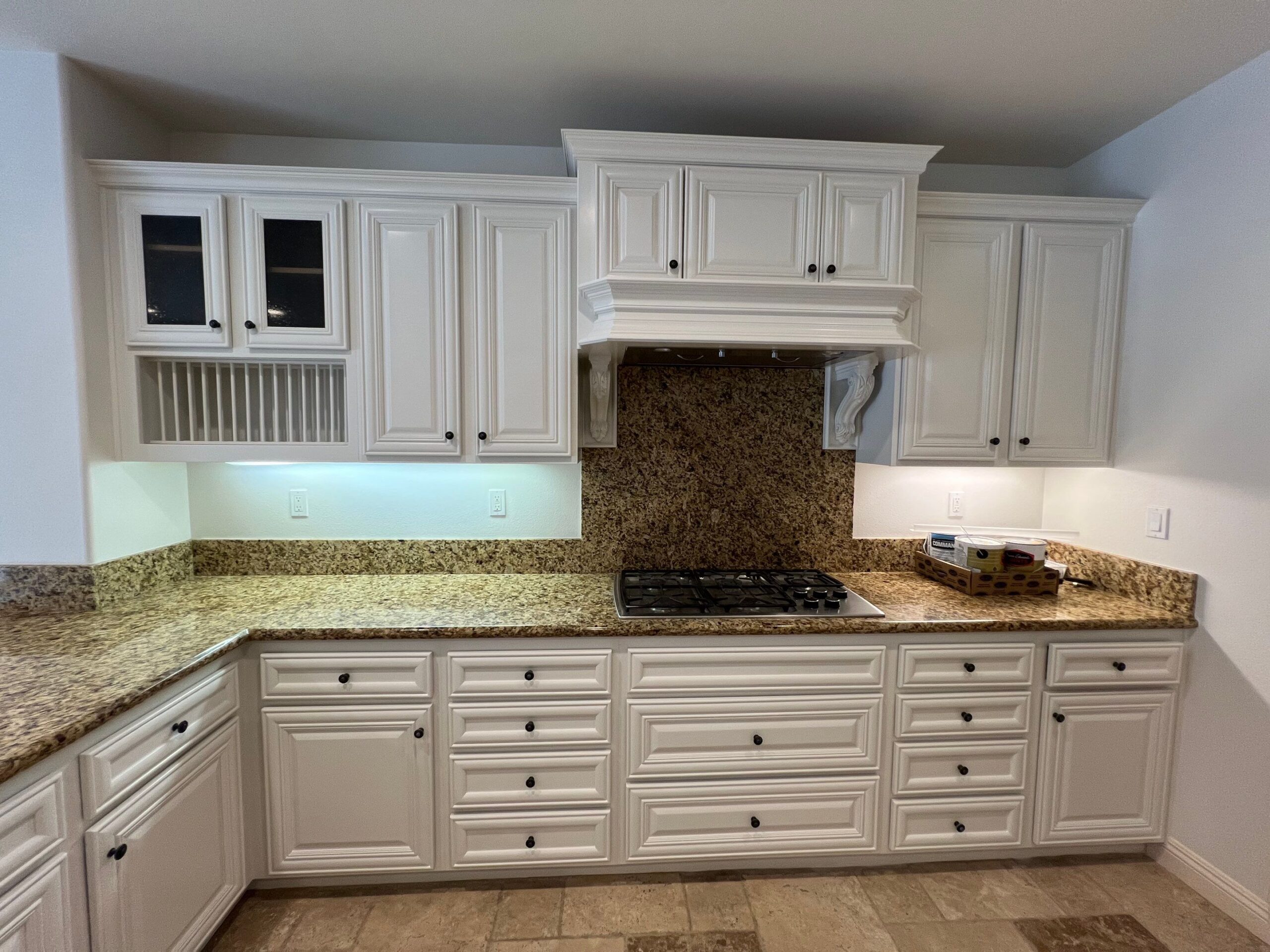 Cream Painted Kitchen Cabinets Cream-painted kitchen cabinets with granite countertops, black gas cooktop, and brown tile flooring in residential interior